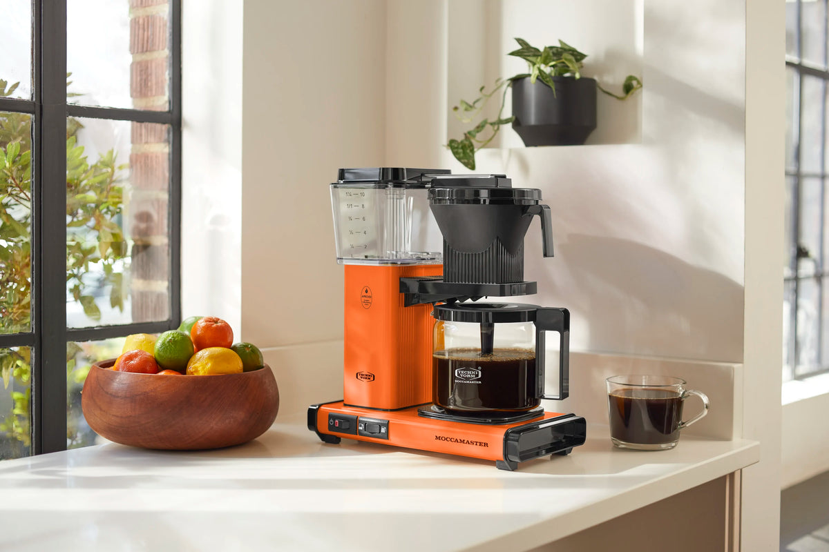 Orange coffee maker on kitchen counter with clear tank and glass carafe beside mug