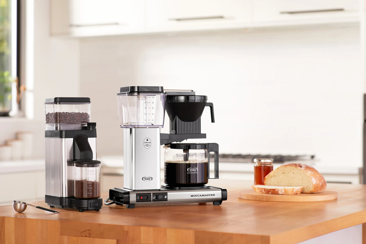 Polished silver burr grinder next to carafe-style coffee maker on wooden counter