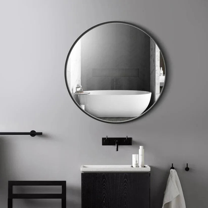 Round wall mirror with black frame reflecting a bathtub and bathroom fixtures