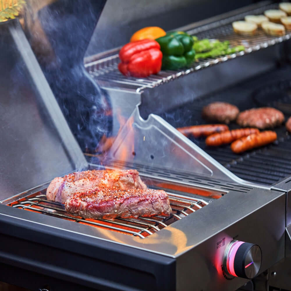 Gas grill with steak on cooking grates, smoke rising, peppers and sausages nearby