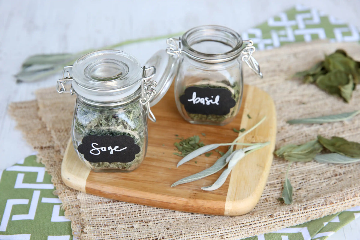Small glass herb jars labeled Sage and Basil on a wooden cutting board