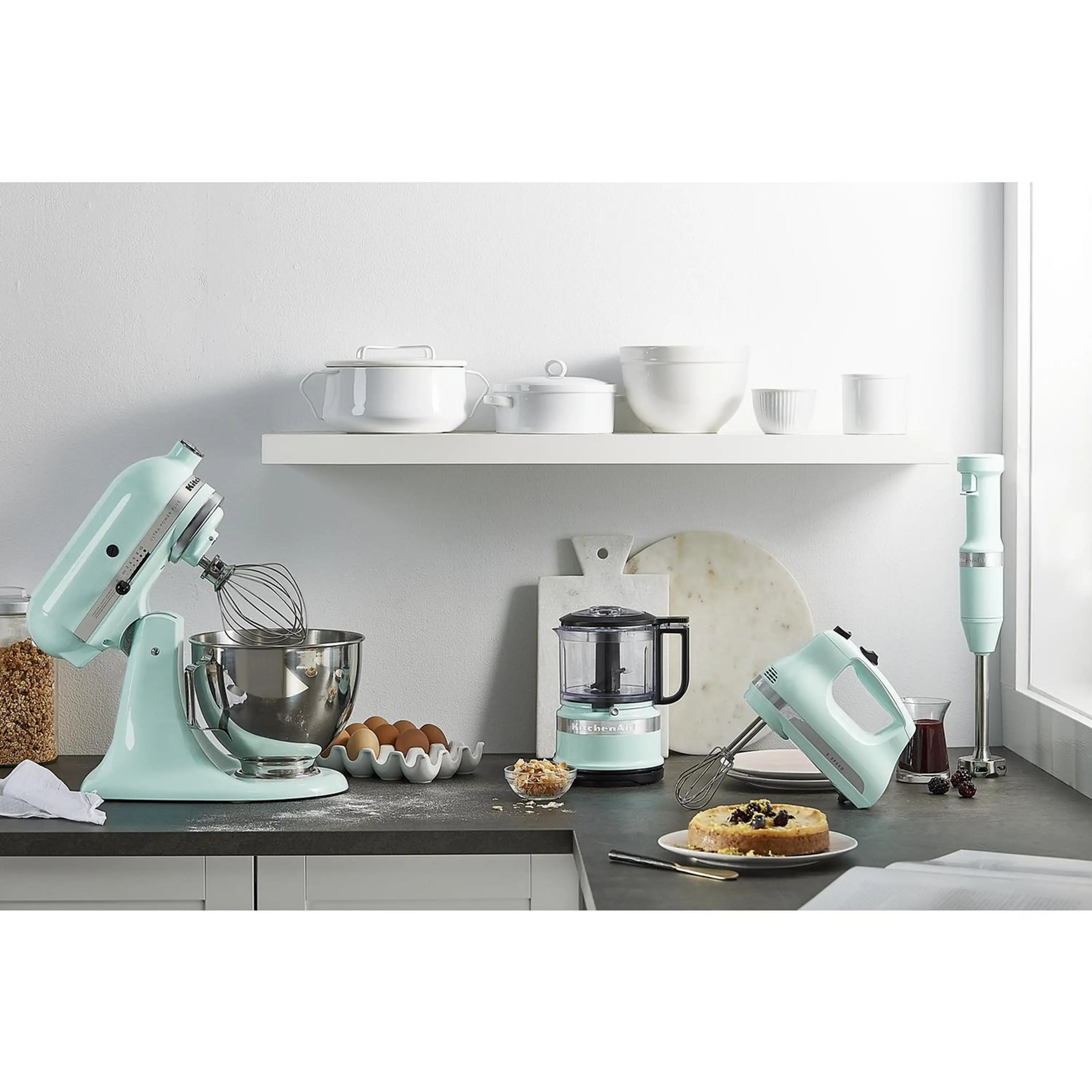 Ice-colored KitchenAid hand mixer with chrome beaters on countertop beside other appliances