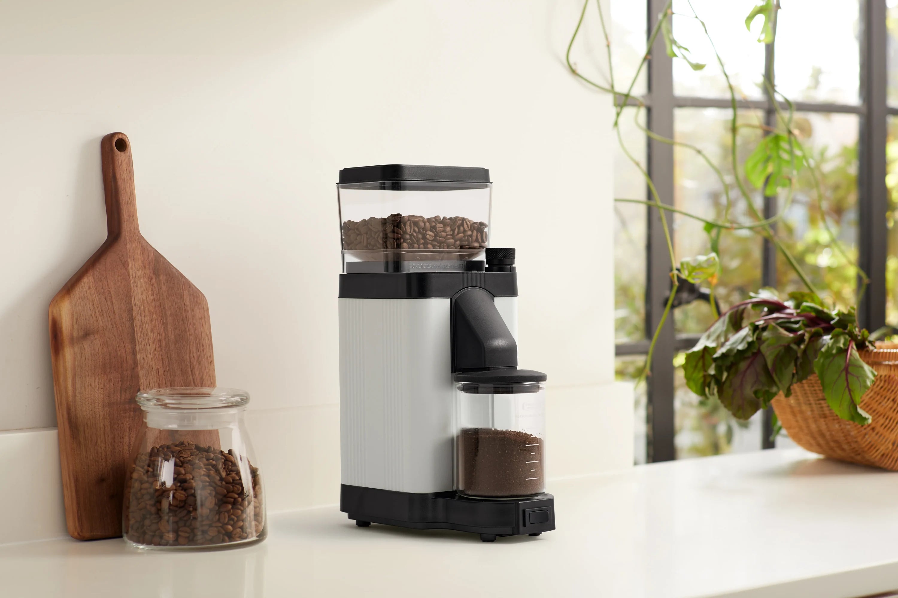 Matte white coffee burr grinder with clear hopper and glass jar of ground coffee on counter.
