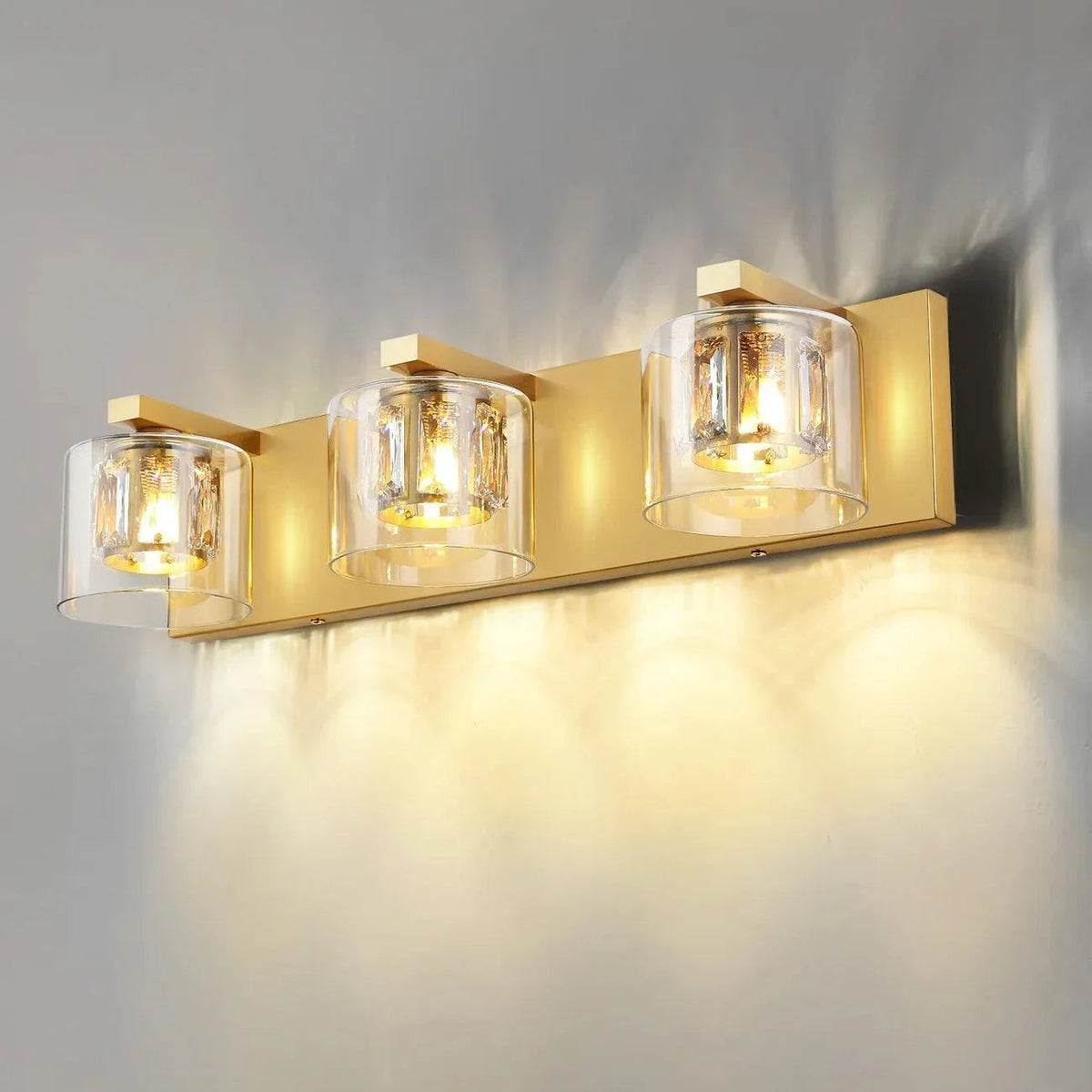 Golden 3-light vanity light with clear glass shades and warm lit bulbs on wall