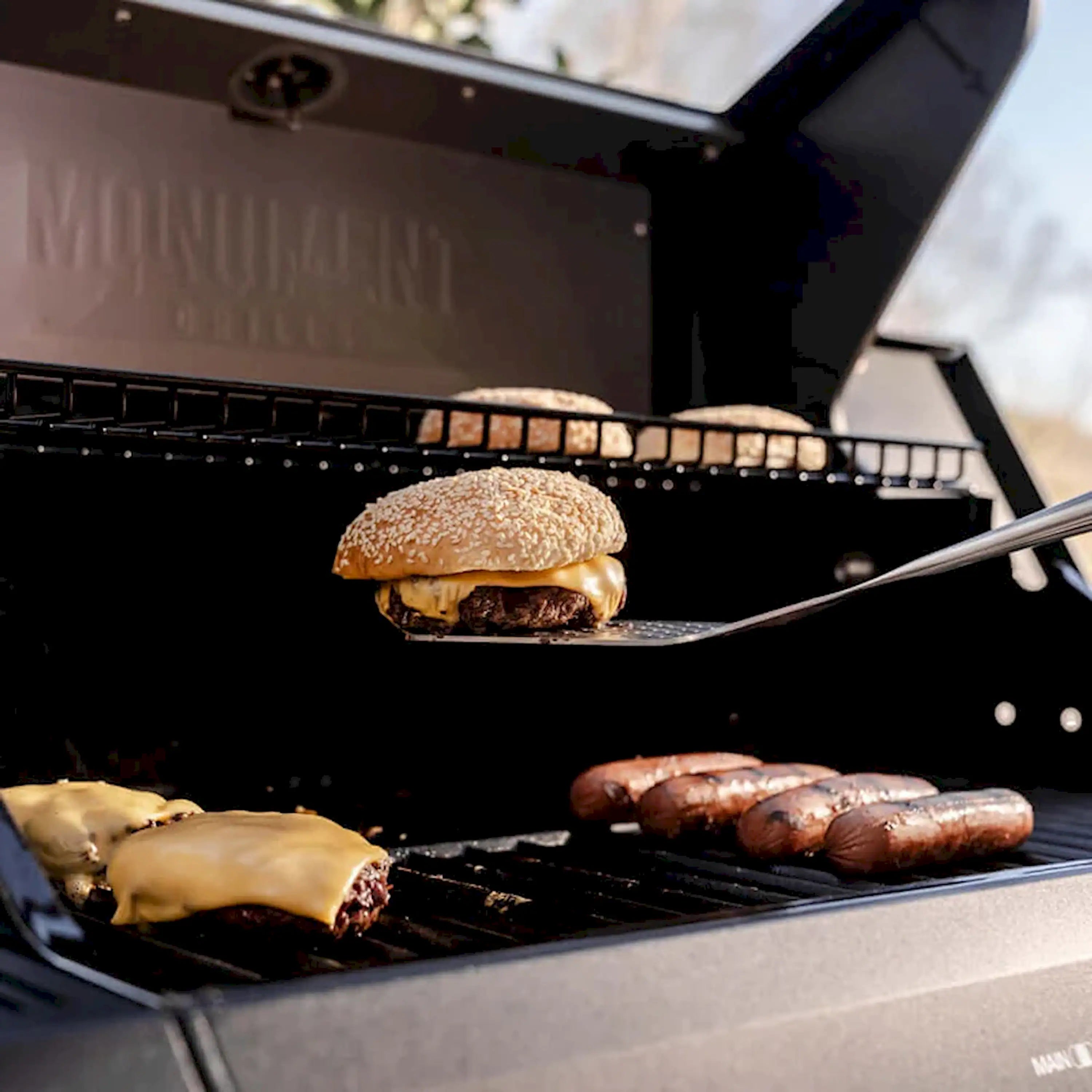 Black propane grill cooking surface with burger and sausages under the open lid