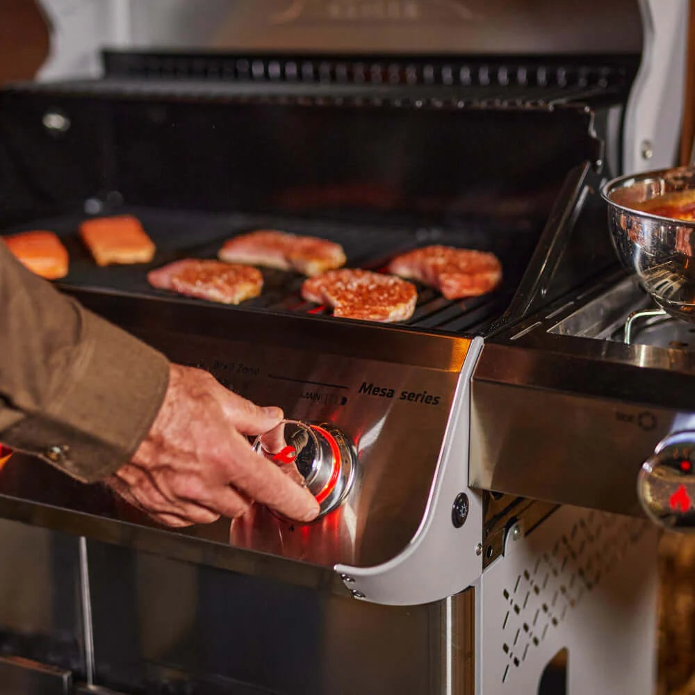 Stainless steel propane gas grill with open lid; hand adjusts burner knobs while cooking food