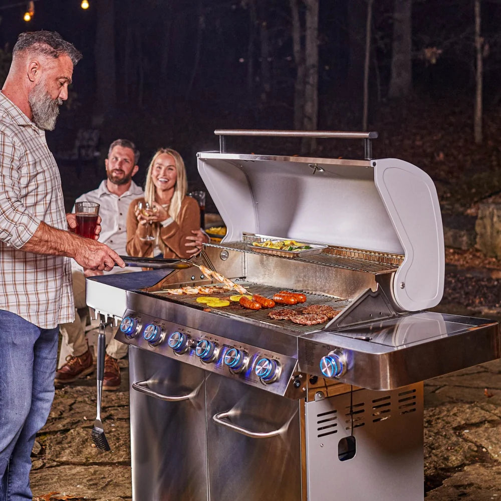 Open stainless propane grill grilling sausages and vegetables with six lit blue burners