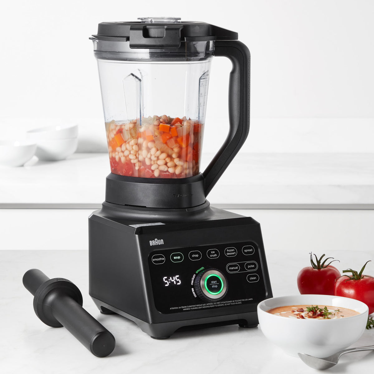 Black blender with clear jar containing red ingredients, on countertop with bowls.