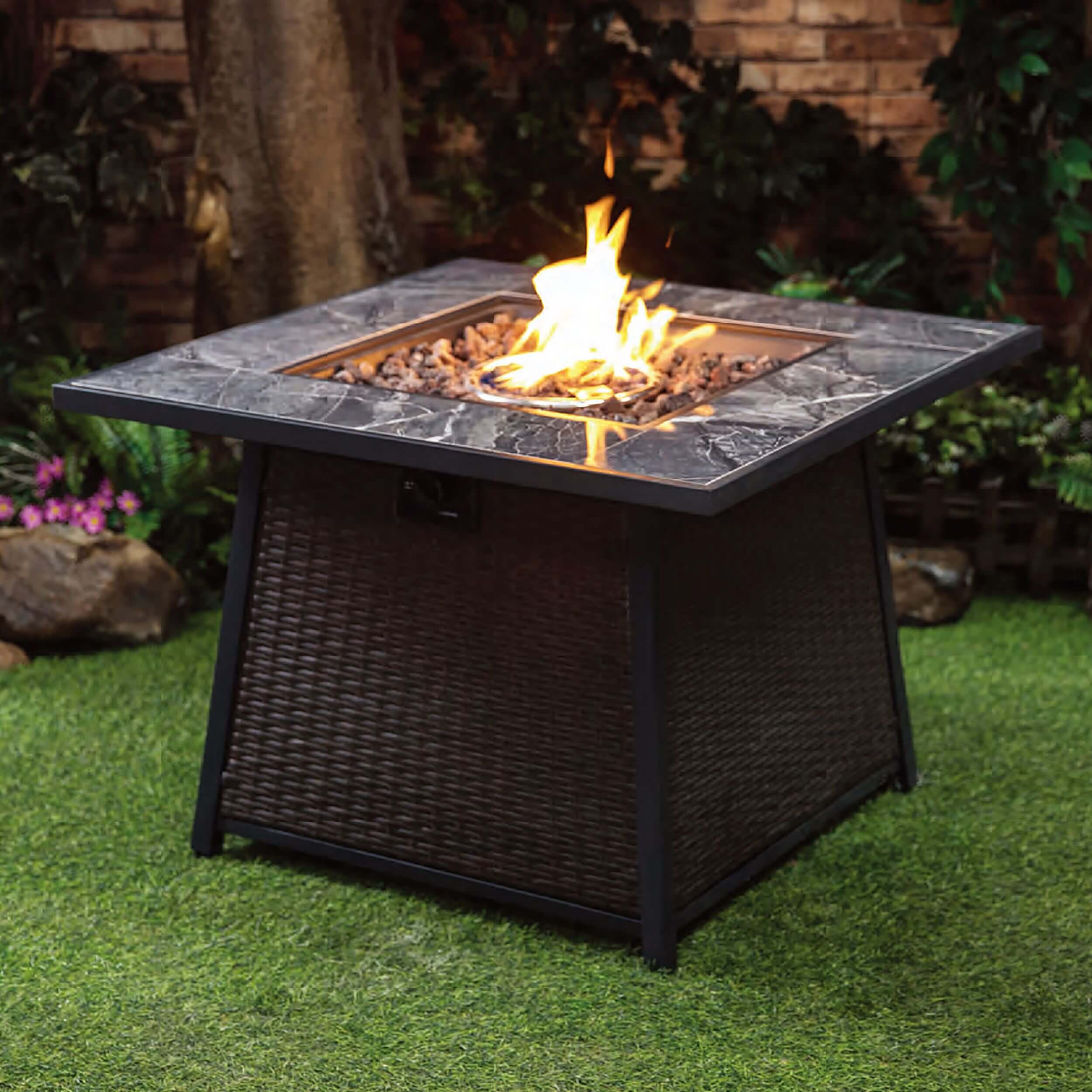 Outdoor gas firepit table with square gray stone top and dark woven base, flames visible