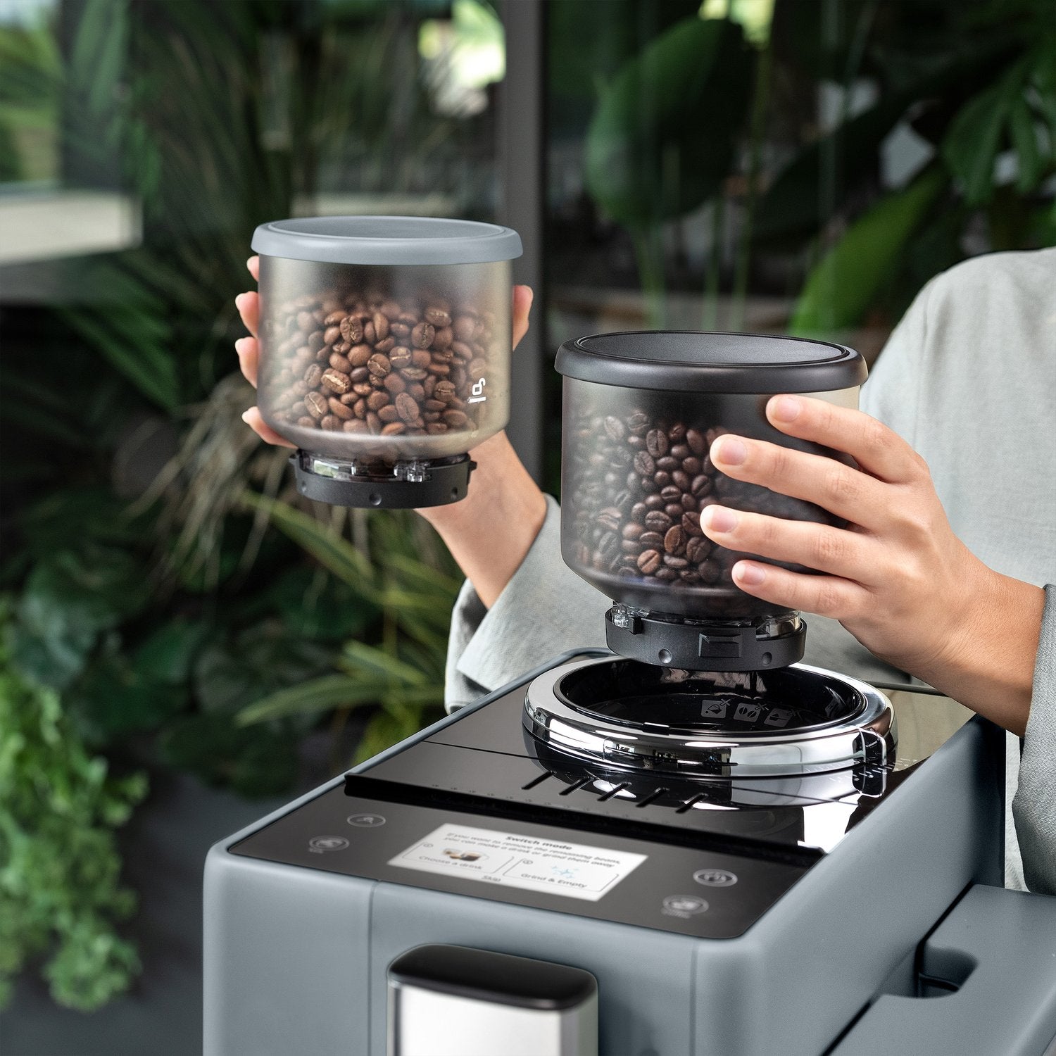 Pebble gray espresso machine with hands holding two bean hoppers over the top station