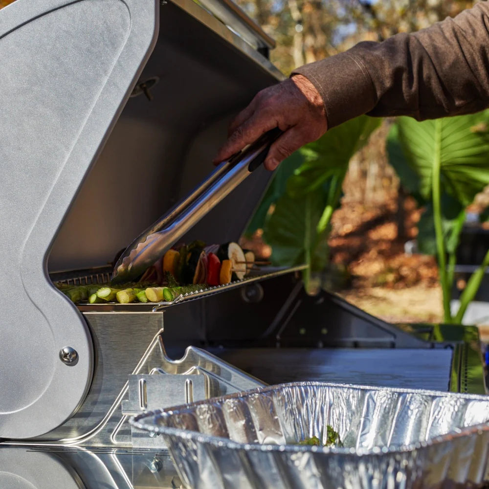 Open stainless steel grill lid with foil tray and hand reaching to place food