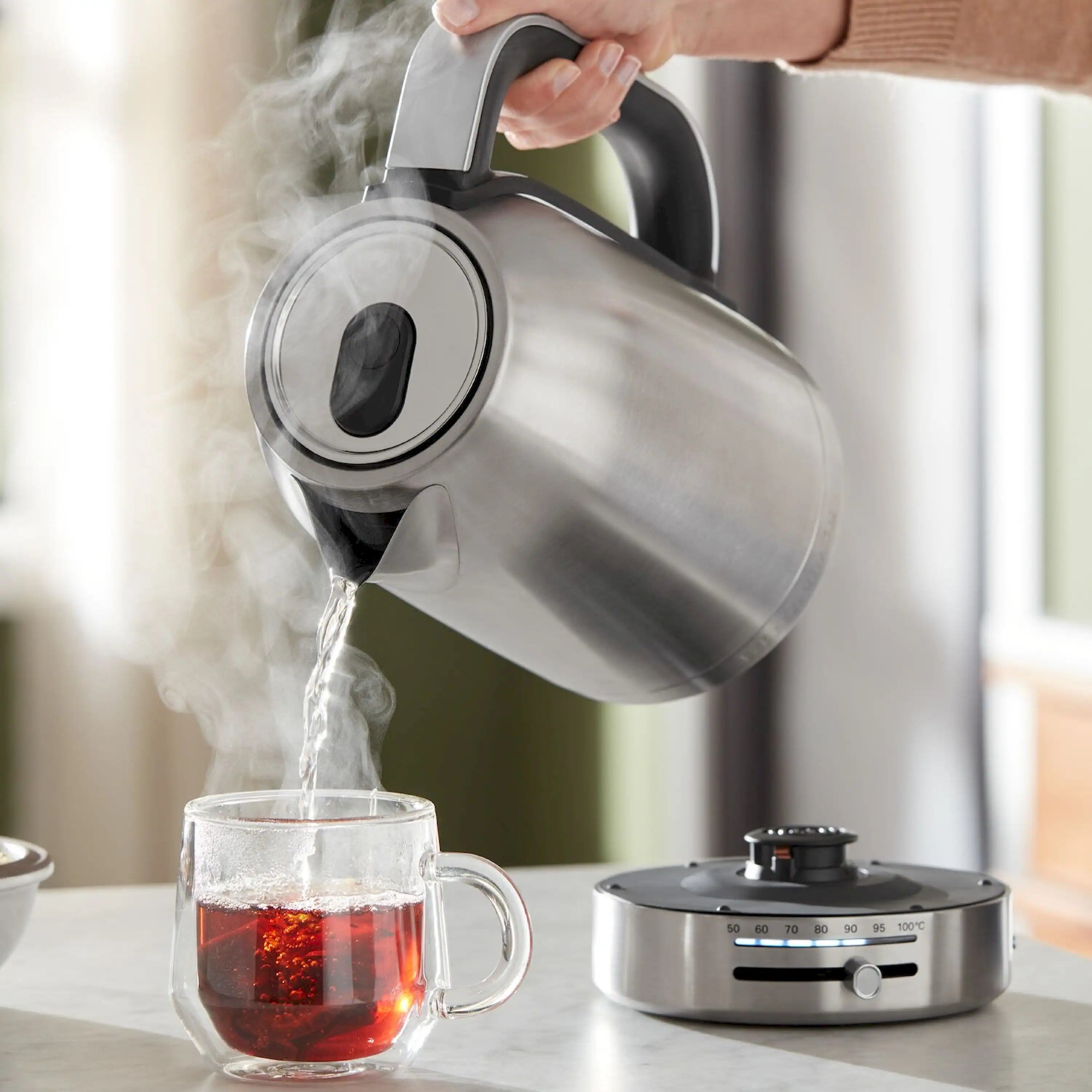 Brushed stainless steel kettle pouring hot water into a glass cup with steam