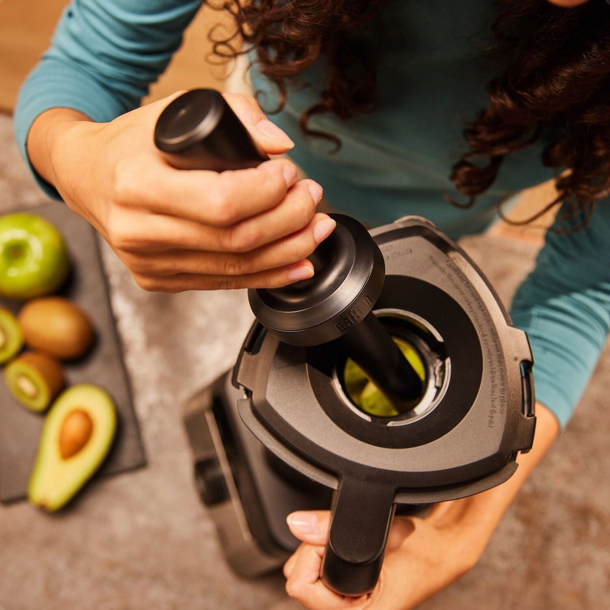 Hand holding black blending attachment over blender jar with avocado slices nearby.