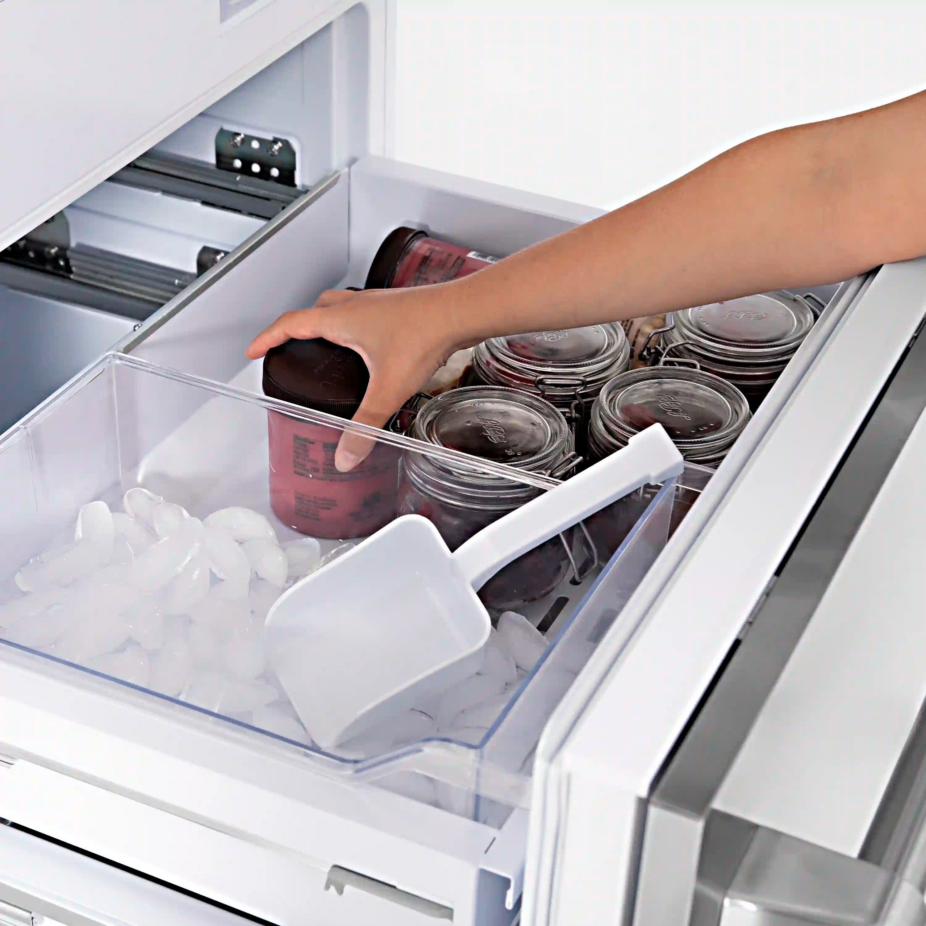 Bottom freezer refrigerator freezer bin with ice tray and hand reaching for stored jars