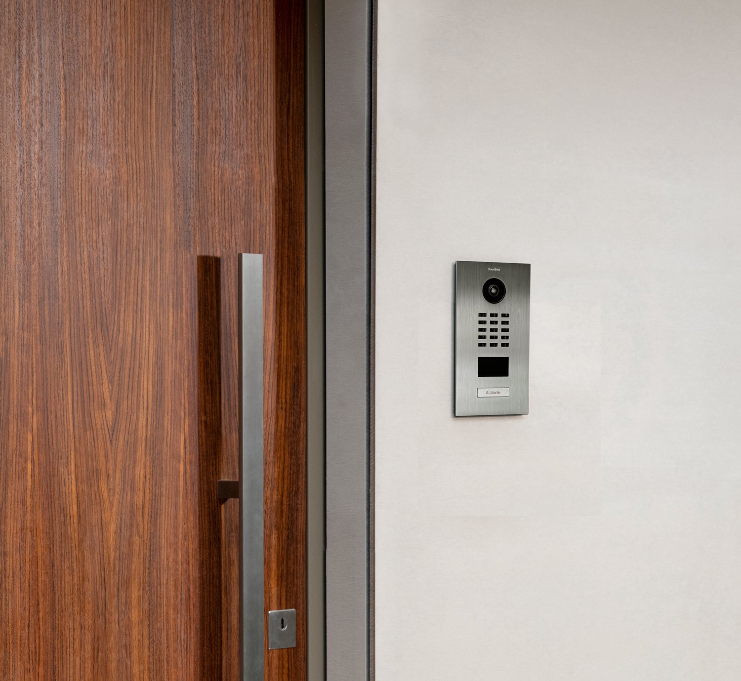Video door station mounted on exterior wall beside wood door frame.