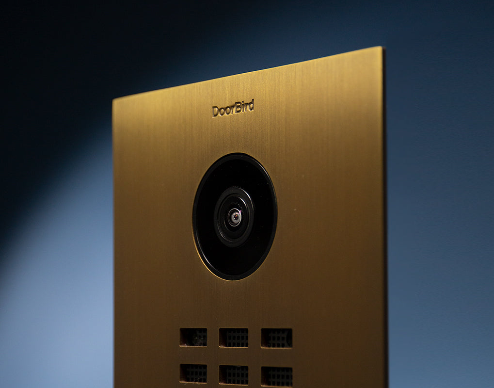 Brass video door station face with centered circular camera and dark speaker ring
