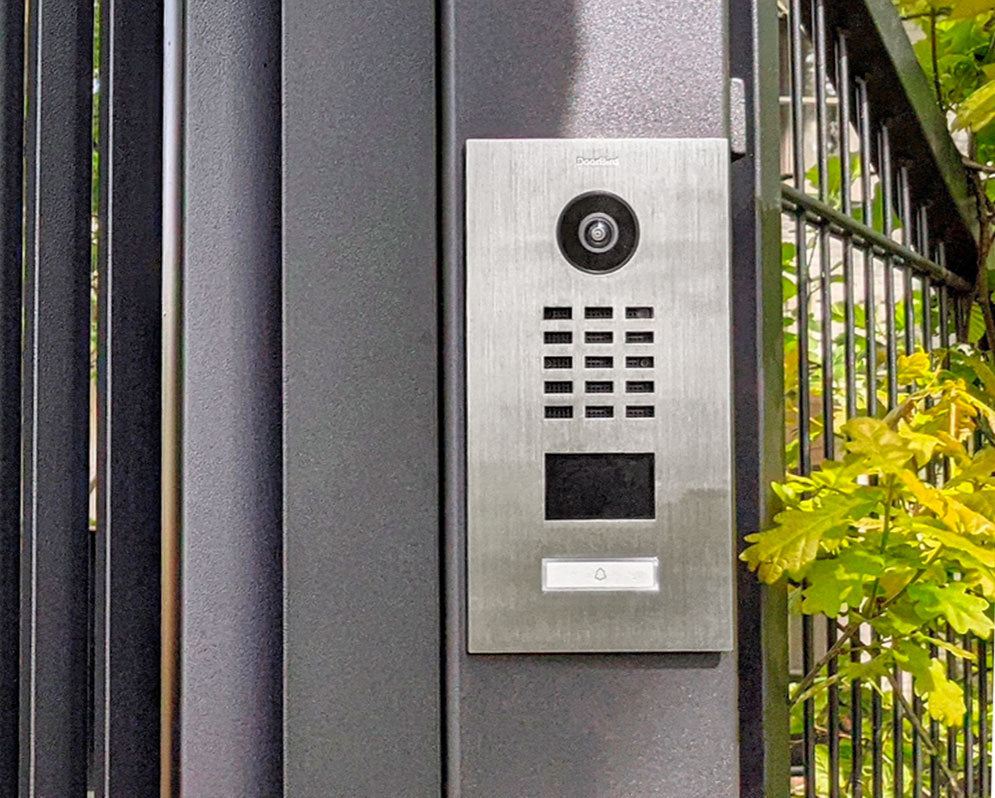 Stainless steel video door station with camera, keypad, and call button near railing.