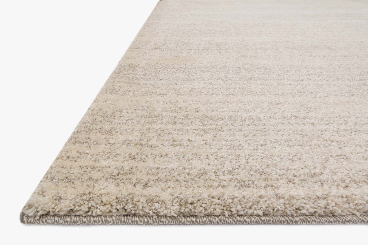 Corner view of speckled light beige power-loomed rug with short fringe edge.