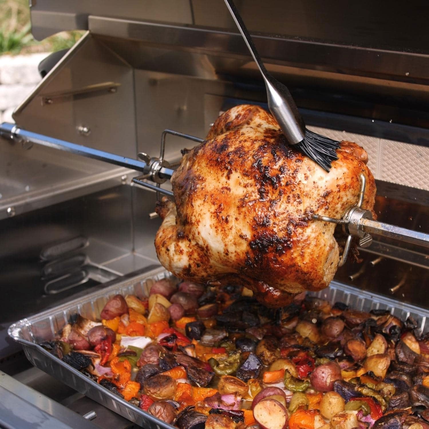 Rotisserie spit with chicken on grill, secured with prongs over roasting tray