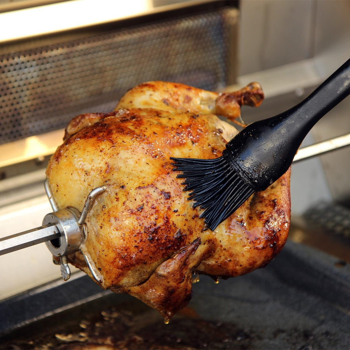 Roasted chicken on rotisserie spit above grill grates with a basting brush