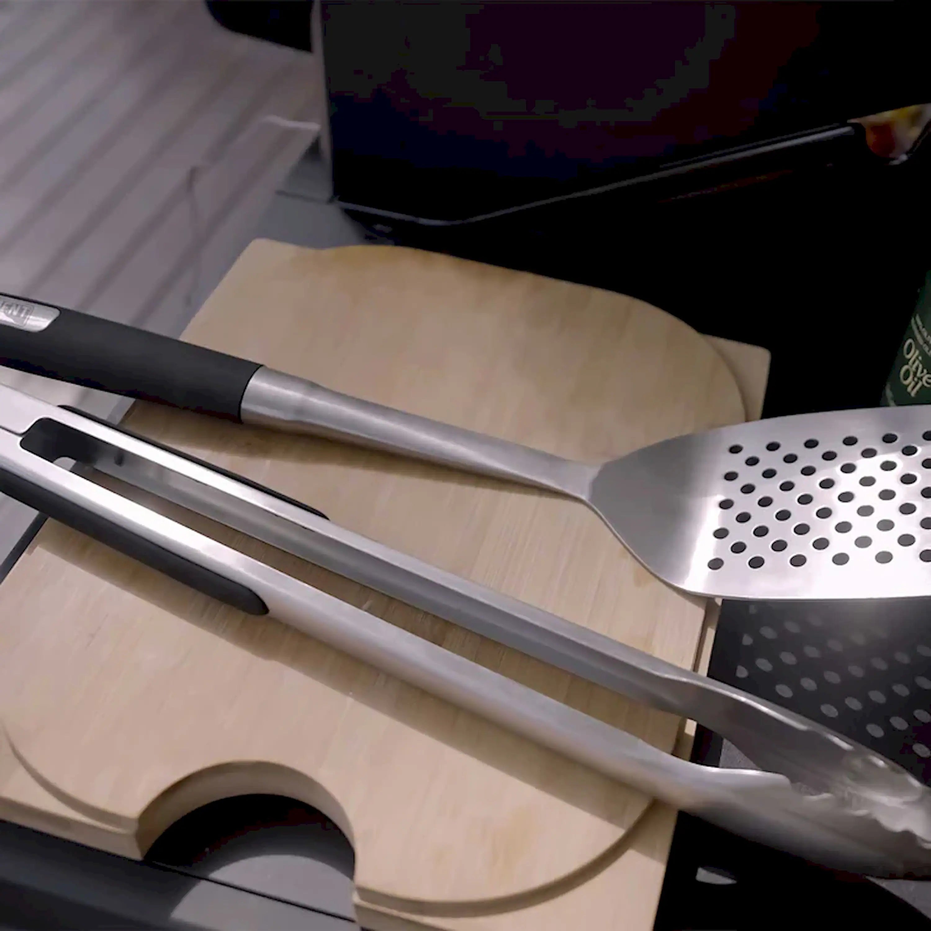 Stainless steel tong and perforated spatula with black handles on wooden board