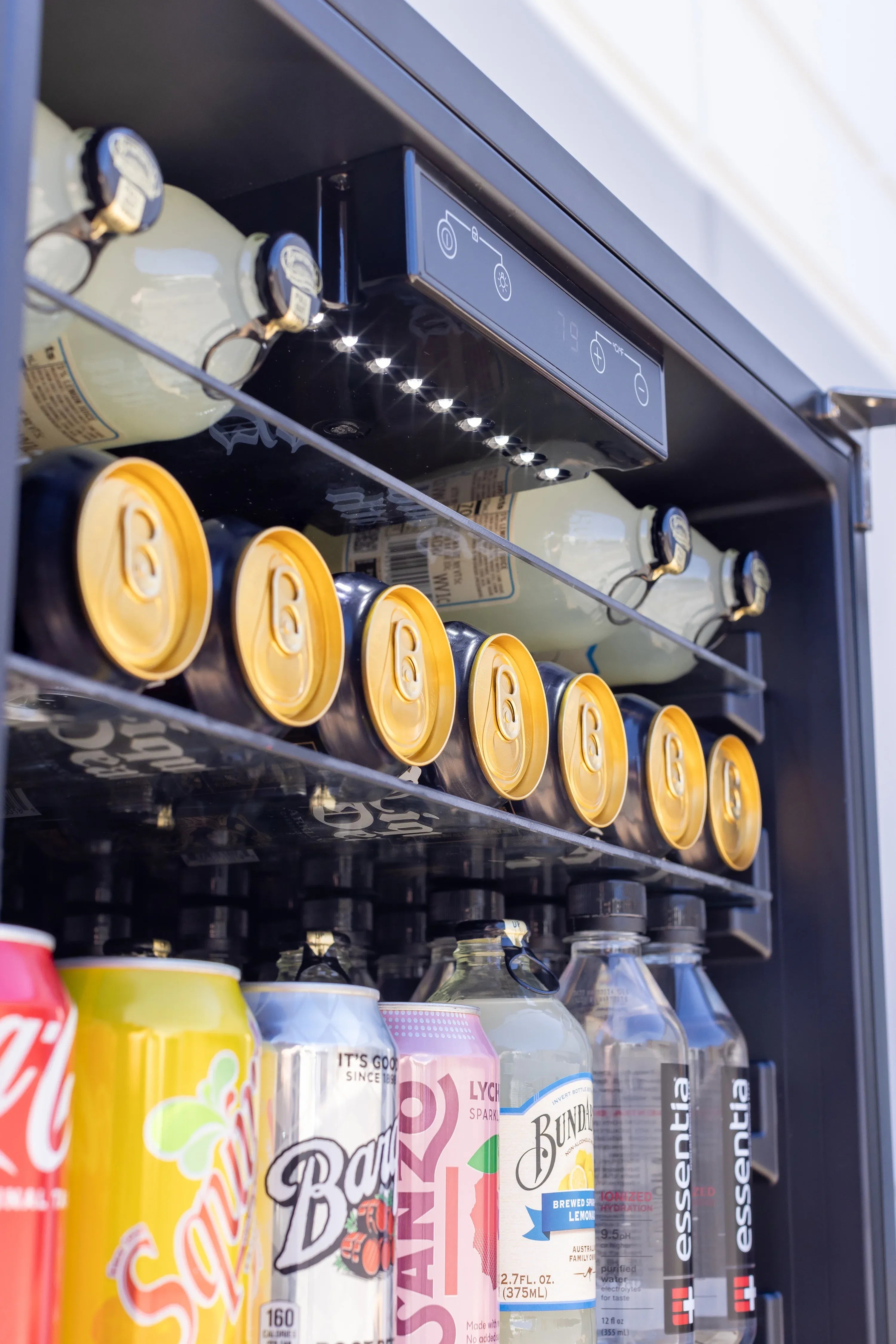 Outdoor fridge interior with lit ceiling panel and multiple drink cans on glass shelf