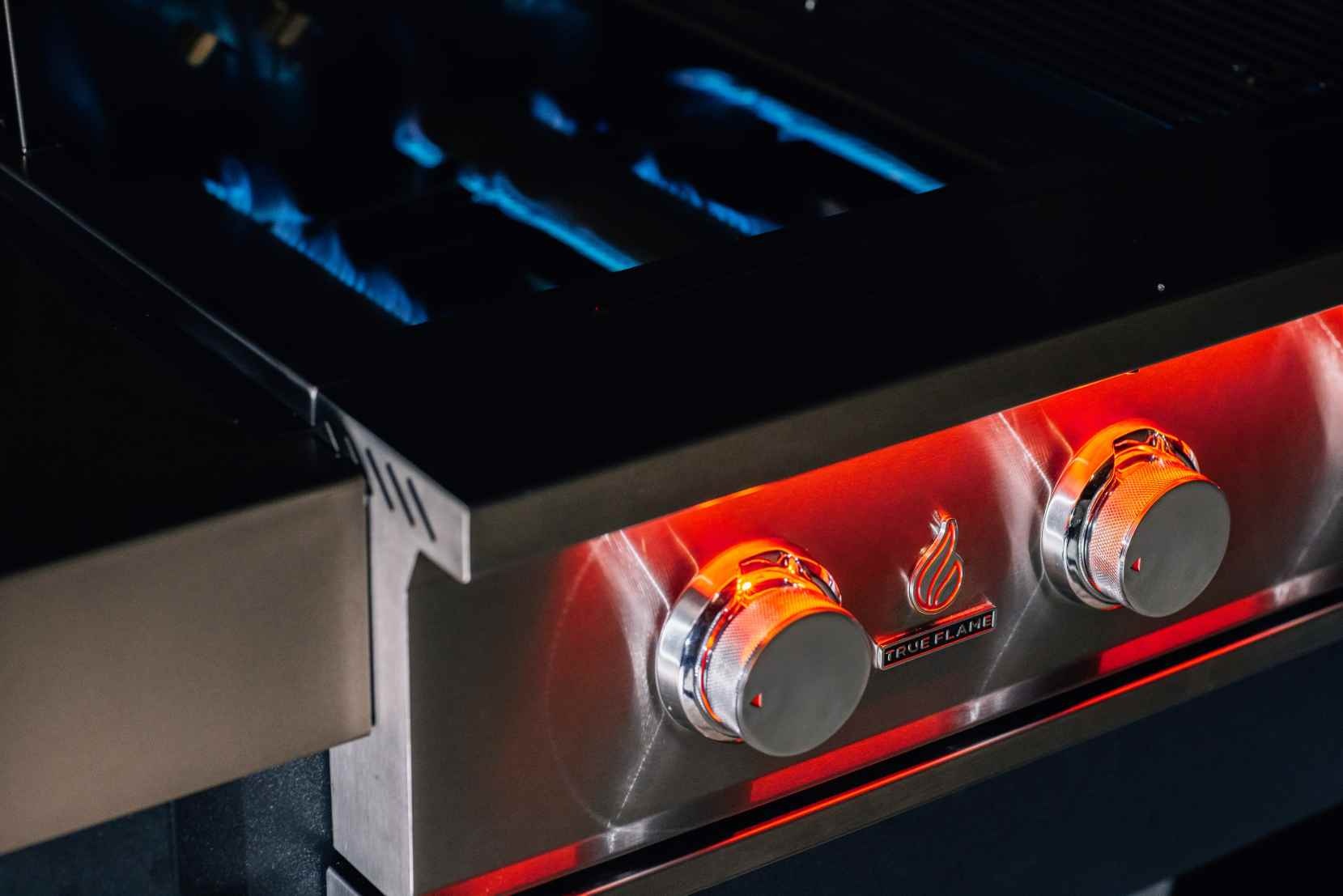Stainless steel gas grill control panel with burner flames behind knobs
