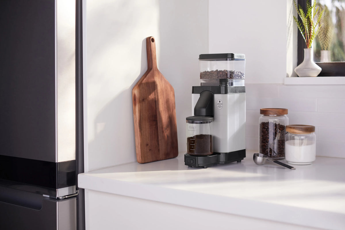Matte white burr grinder on counter with glass jars of beans and ground coffee, side view.