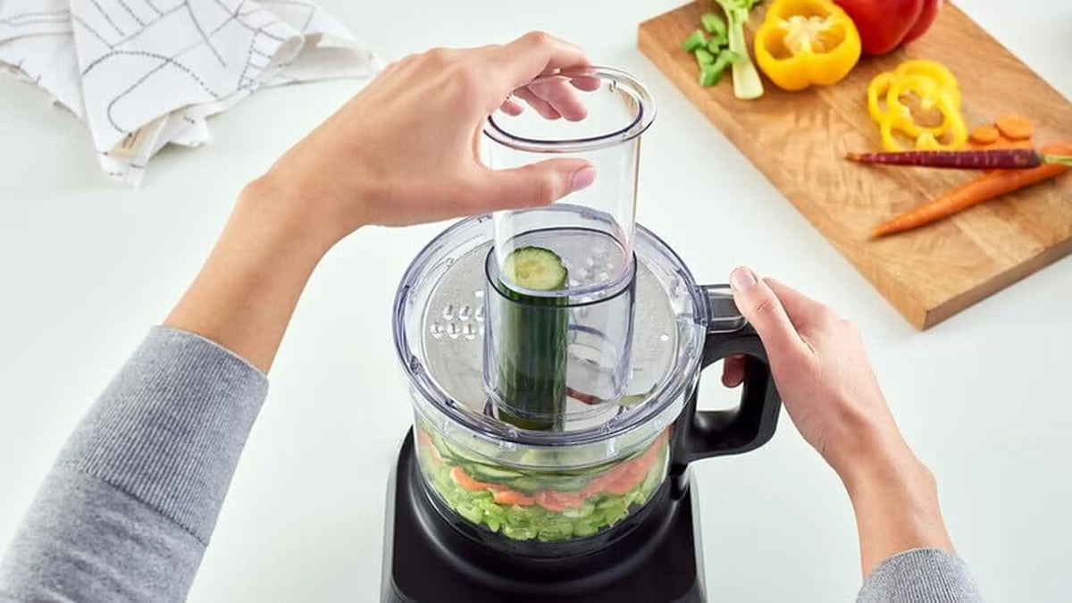 Braun food processor with black base and clear bowl, hands loading cucumber