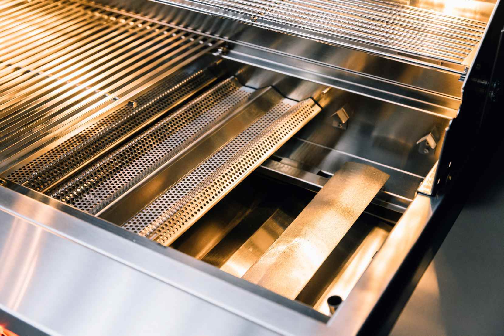 Open stainless steel gas grill firebox with burner tiles and metal heat shield