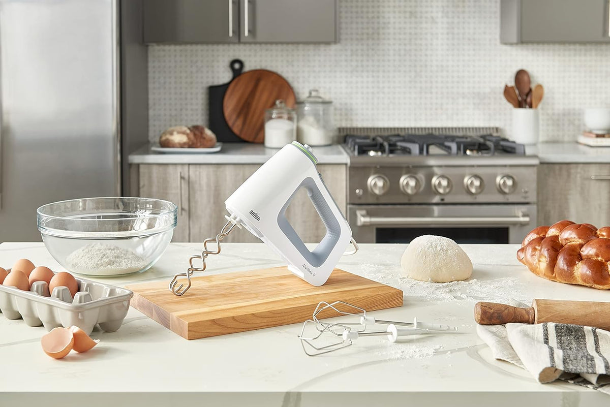 White hand mixer with beaters on kitchen counter with dough and eggs visible