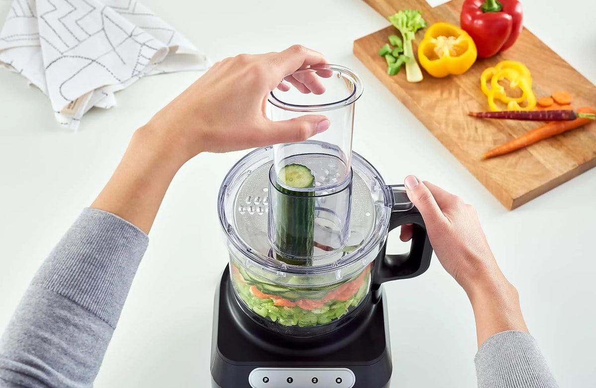 Braun food processor with clear bowl and black base, hands adding chopped vegetables