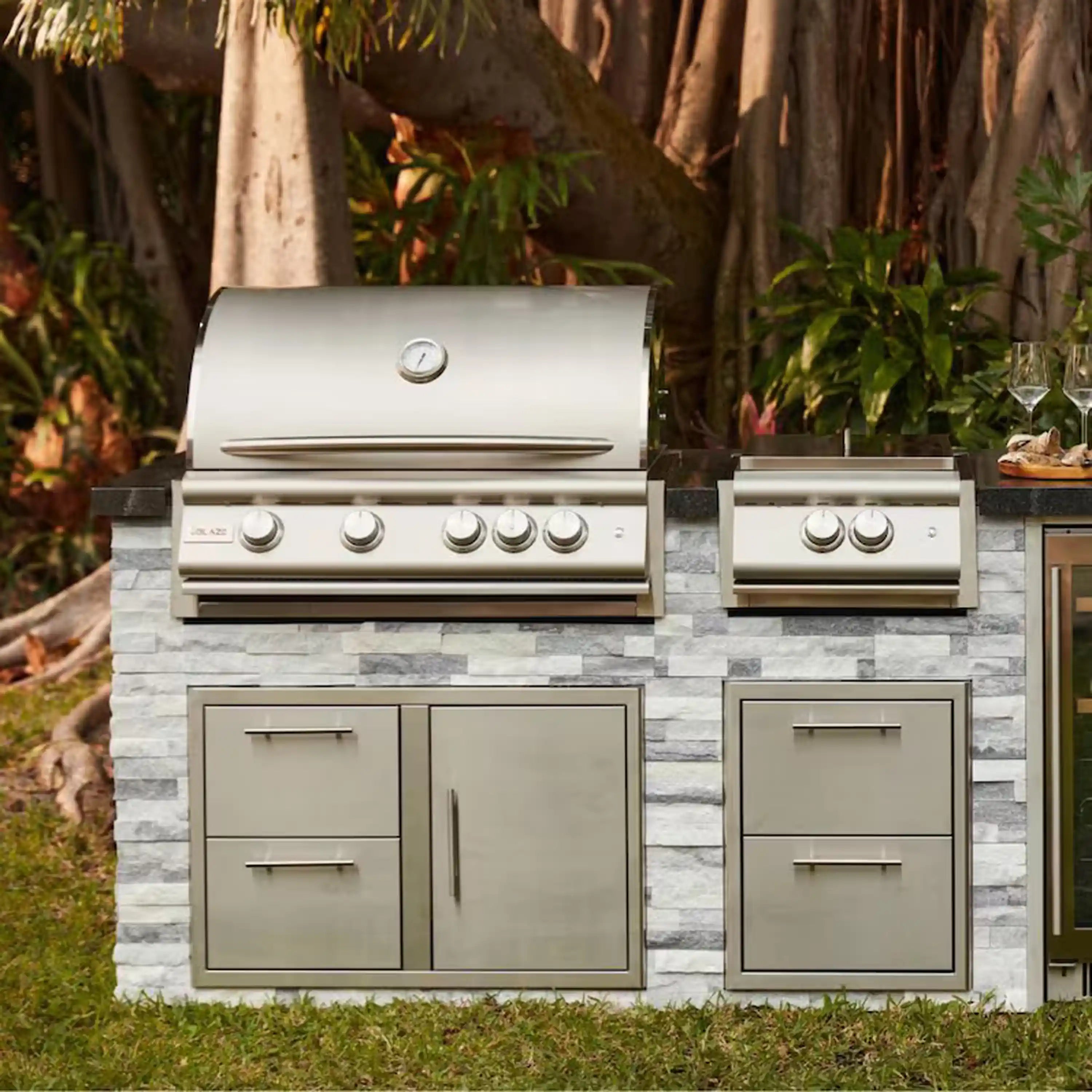 Outdoor built-in gas grill with stainless hood, multiple front control knobs, and drawers