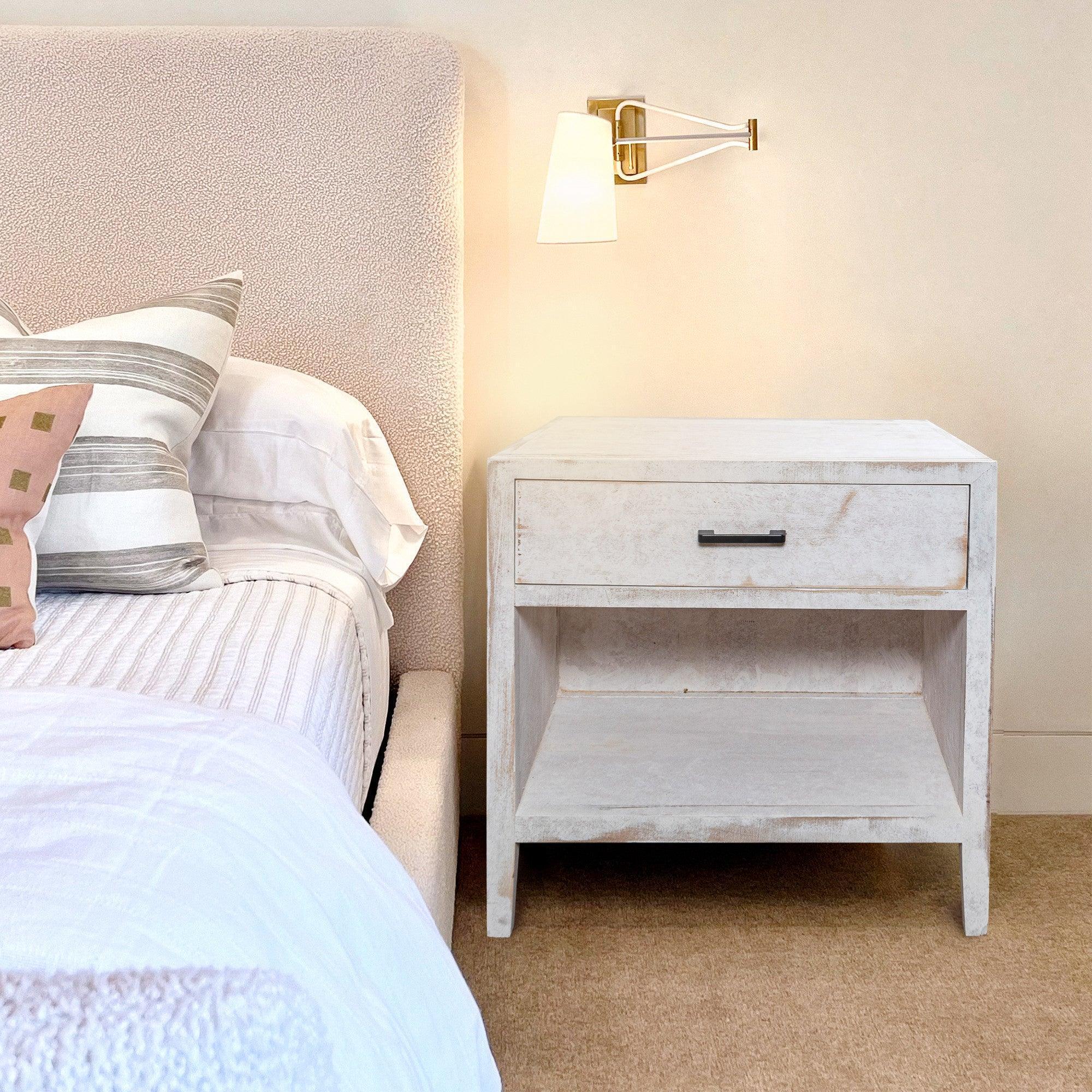 White solid wood nightstand beside bed with single drawer and shelf