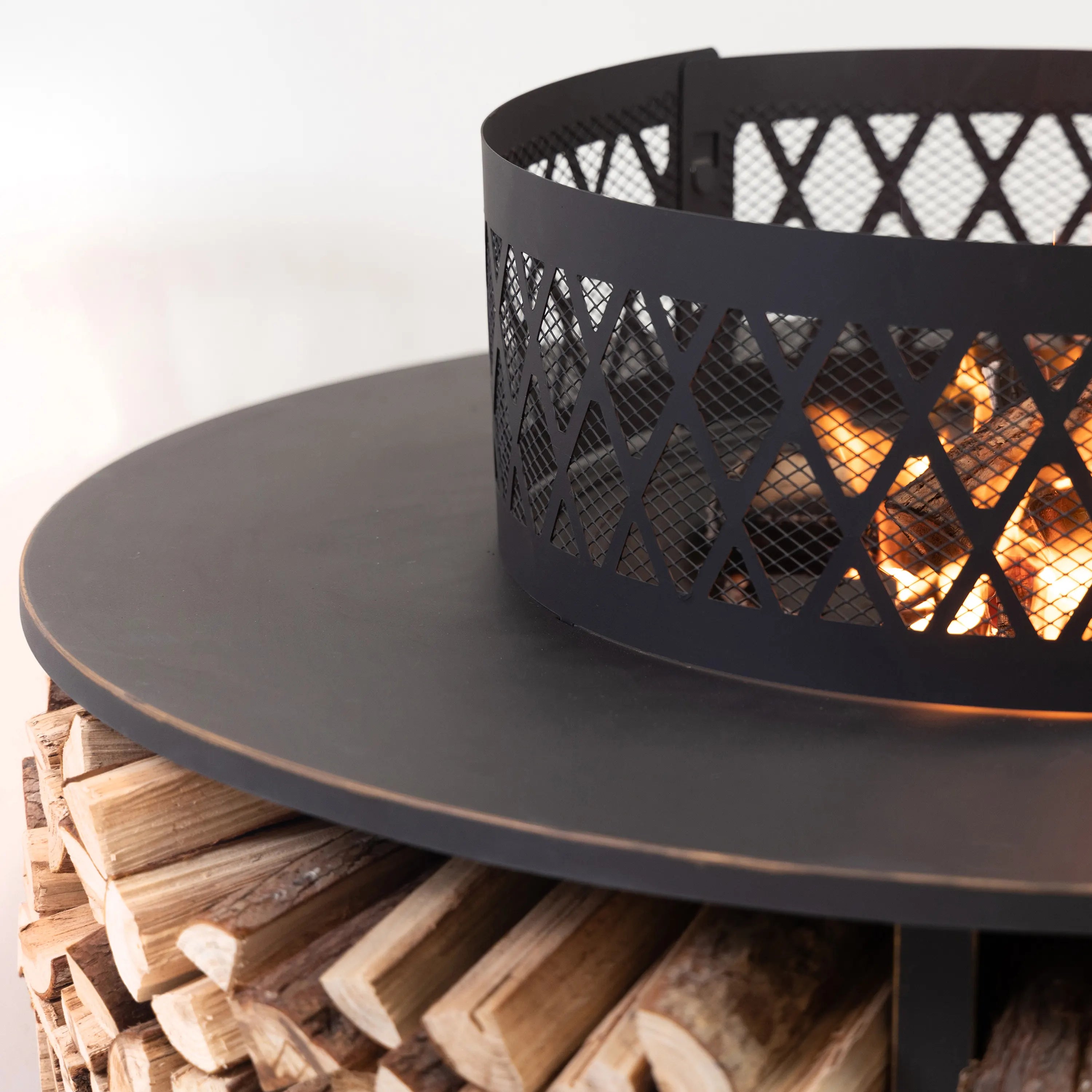 Black mesh fire pit bowl with visible flames and round tabletop over log storage.