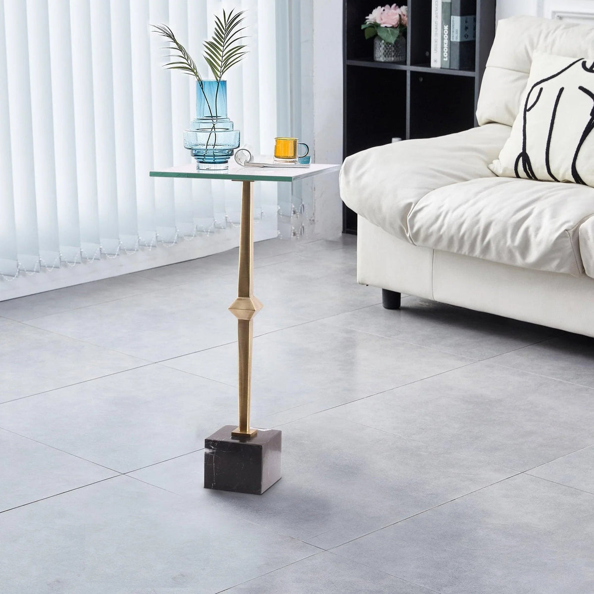 Square side table with glass top, golden leg, and dark marble base beside sofa
