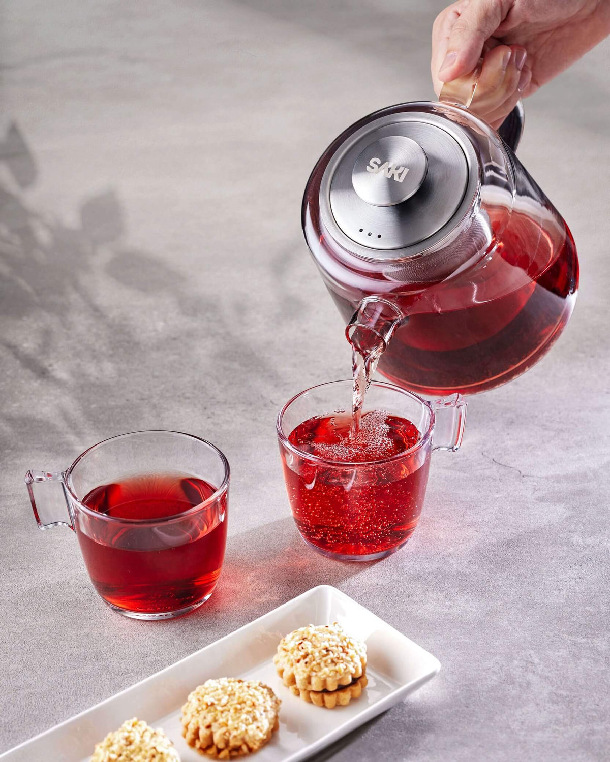 Electric tea maker beside glass kettle pouring red tea into clear cups.