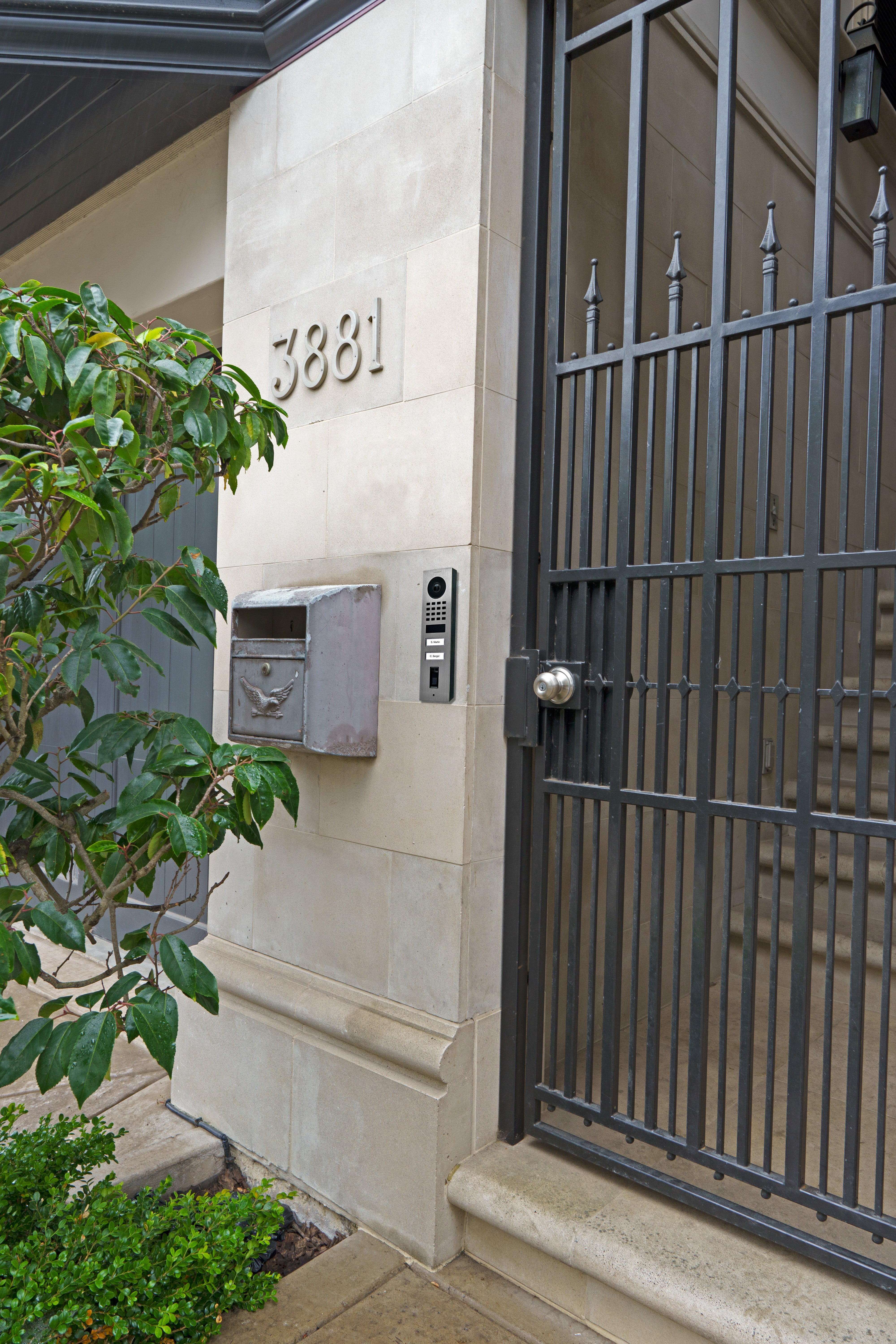 Surface-mounted stainless steel video door intercom on exterior wall next to a gate