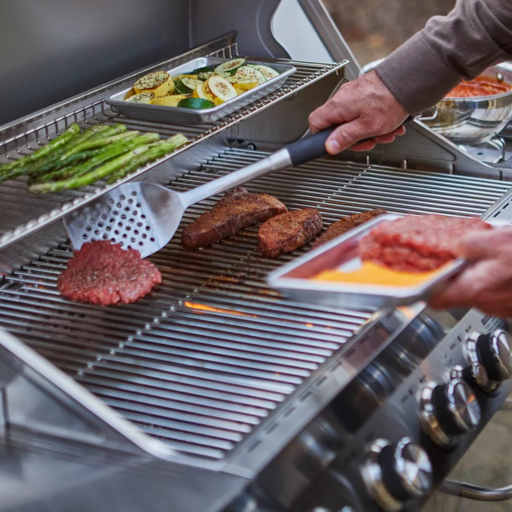 Open stainless steel grill with burgers cooking on grates and utensils in hand