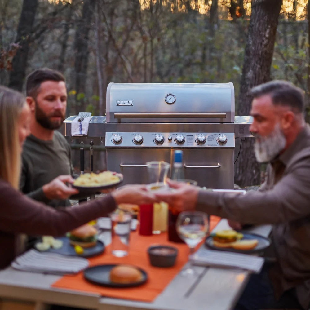 Stainless steel propane gas grill in use behind a table with plated food