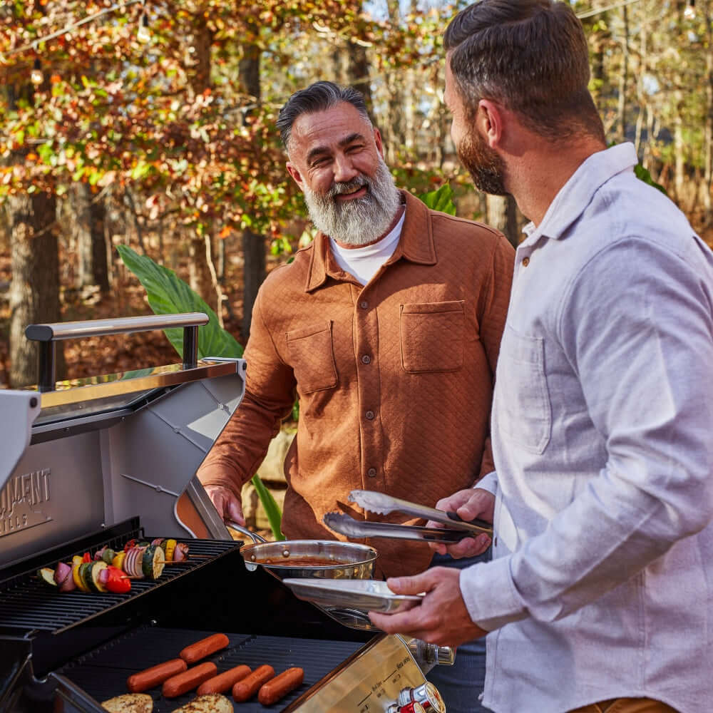 Stainless steel gas grill with lid open; sausages and pan cooking on grates