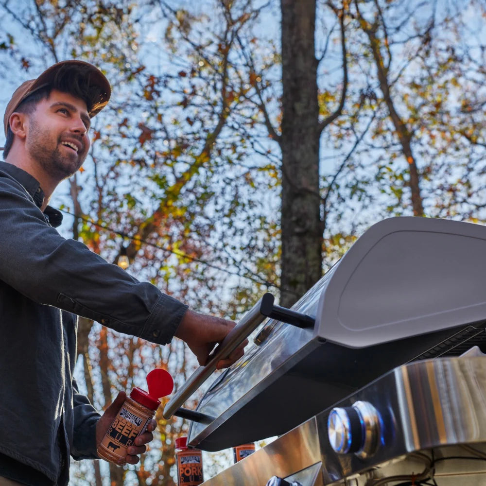 Person cooking at stainless propane grill with open lid and six blue control knobs
