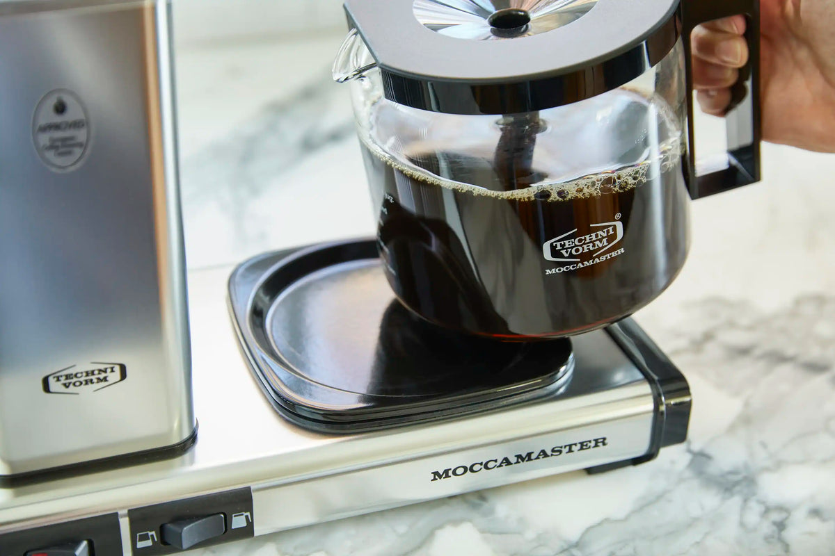 Close-up of Moccamaster glass carafe under brewer, with drip-stop on black base.