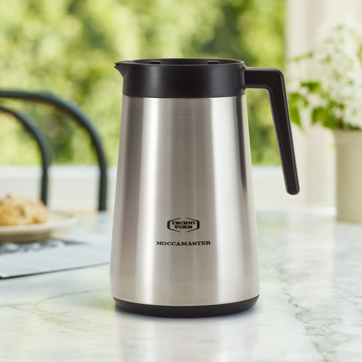 Stainless thermal coffee carafe with black lid and side handle on table