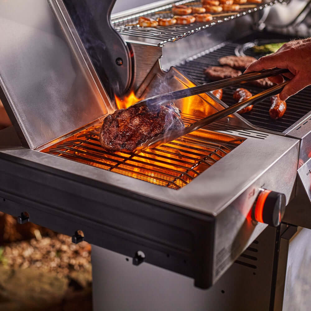 Stainless steel grill grate with searing meat and orange flames beneath the lid