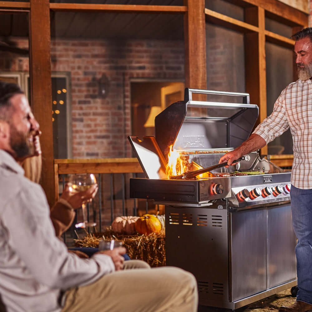 Stainless steel gas grill with open hood, lit burners, and side cooking area