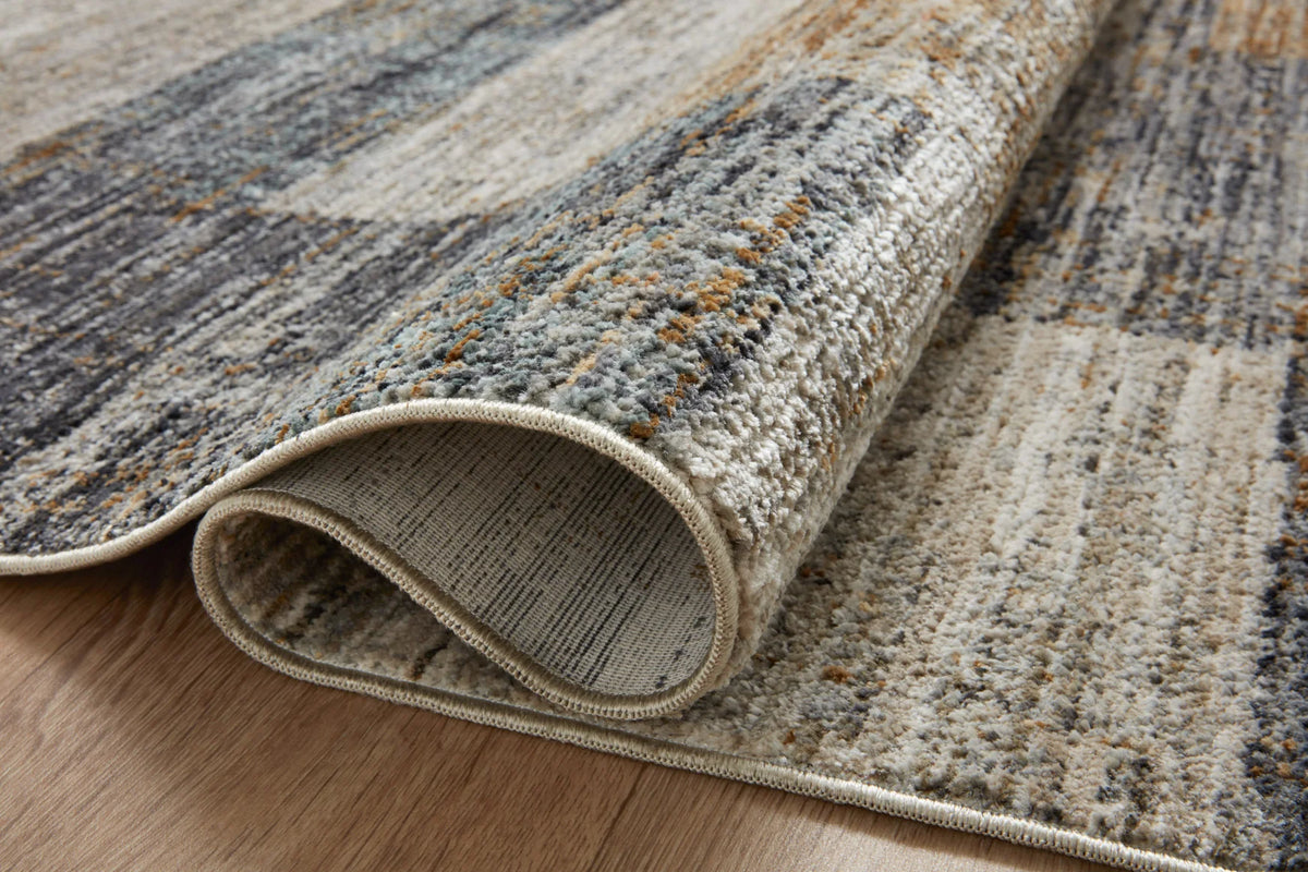 Rolled edge of grey and gold abstract rug with taupe backing visible