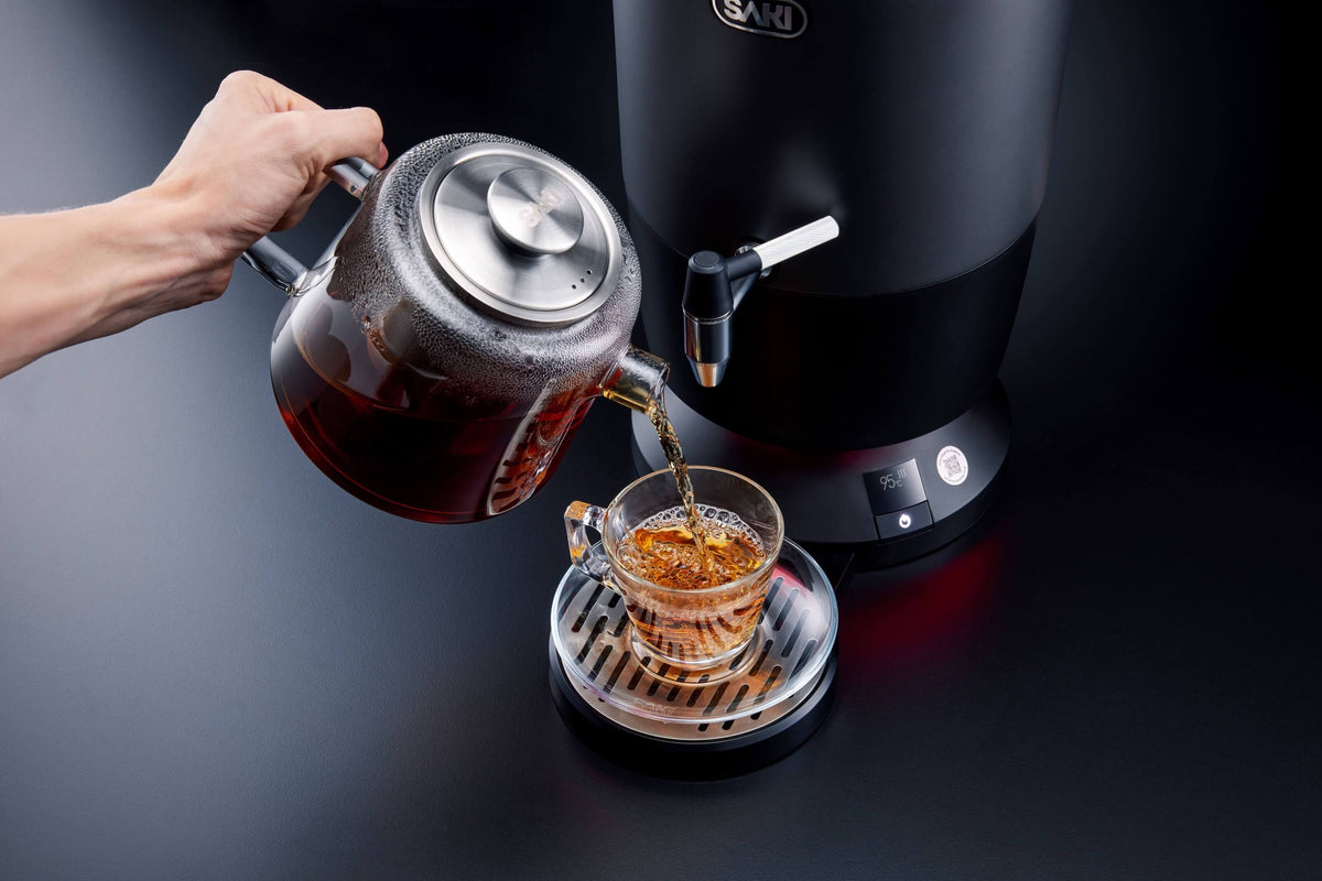 Black electric tea maker with spout and warming basket, pouring hot tea into cups.