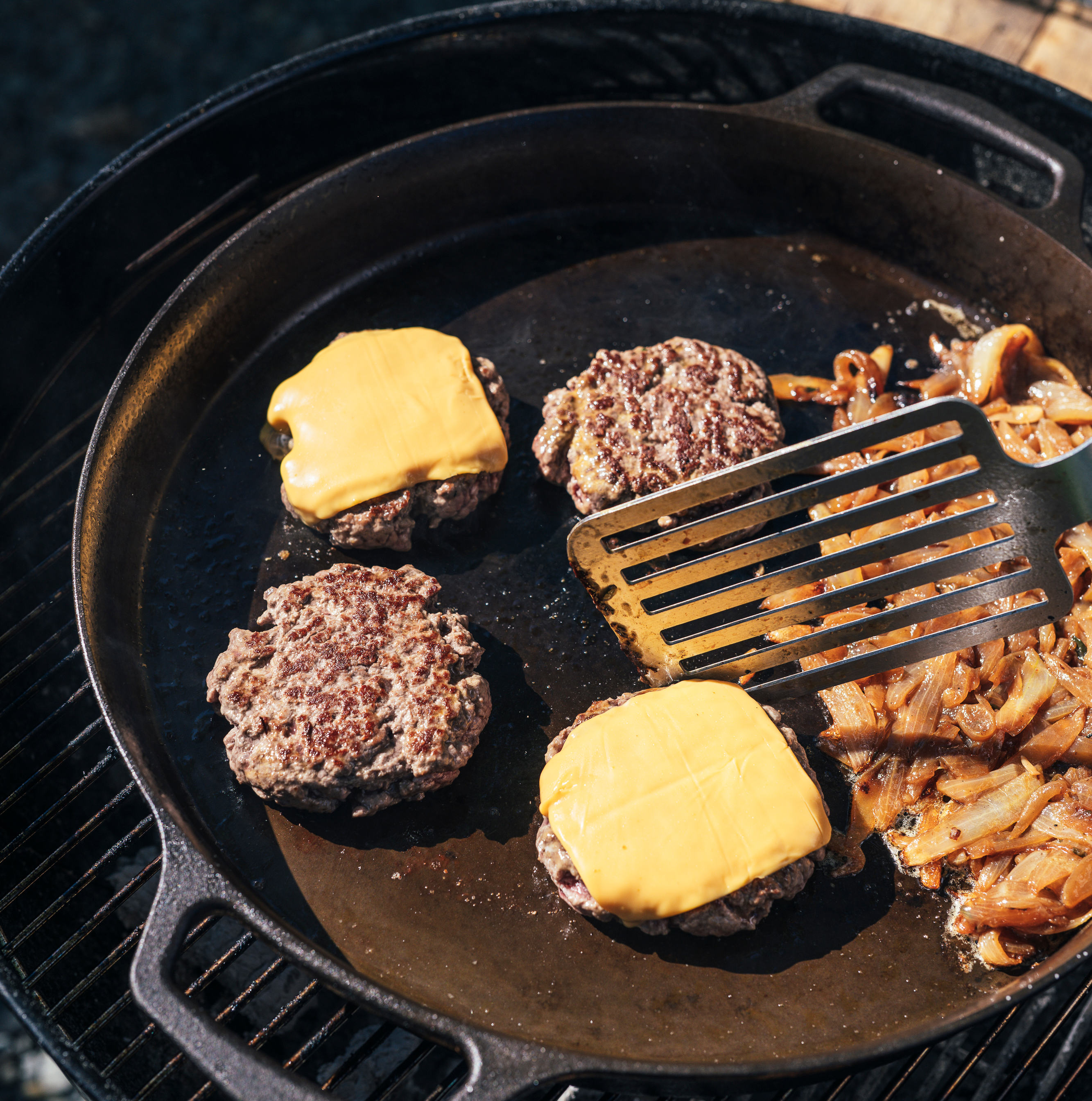 Cast iron skillet on grill grate with burger patties, butter, and onions