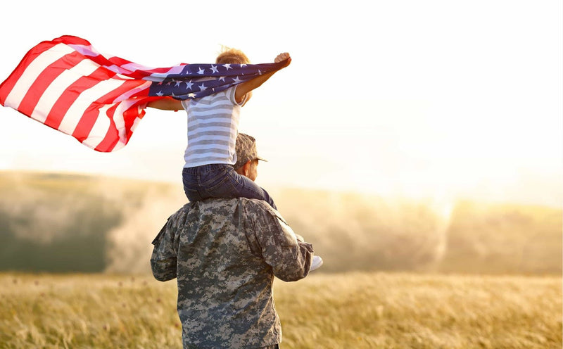 Service member carrying a child on his shoulders under an American flag